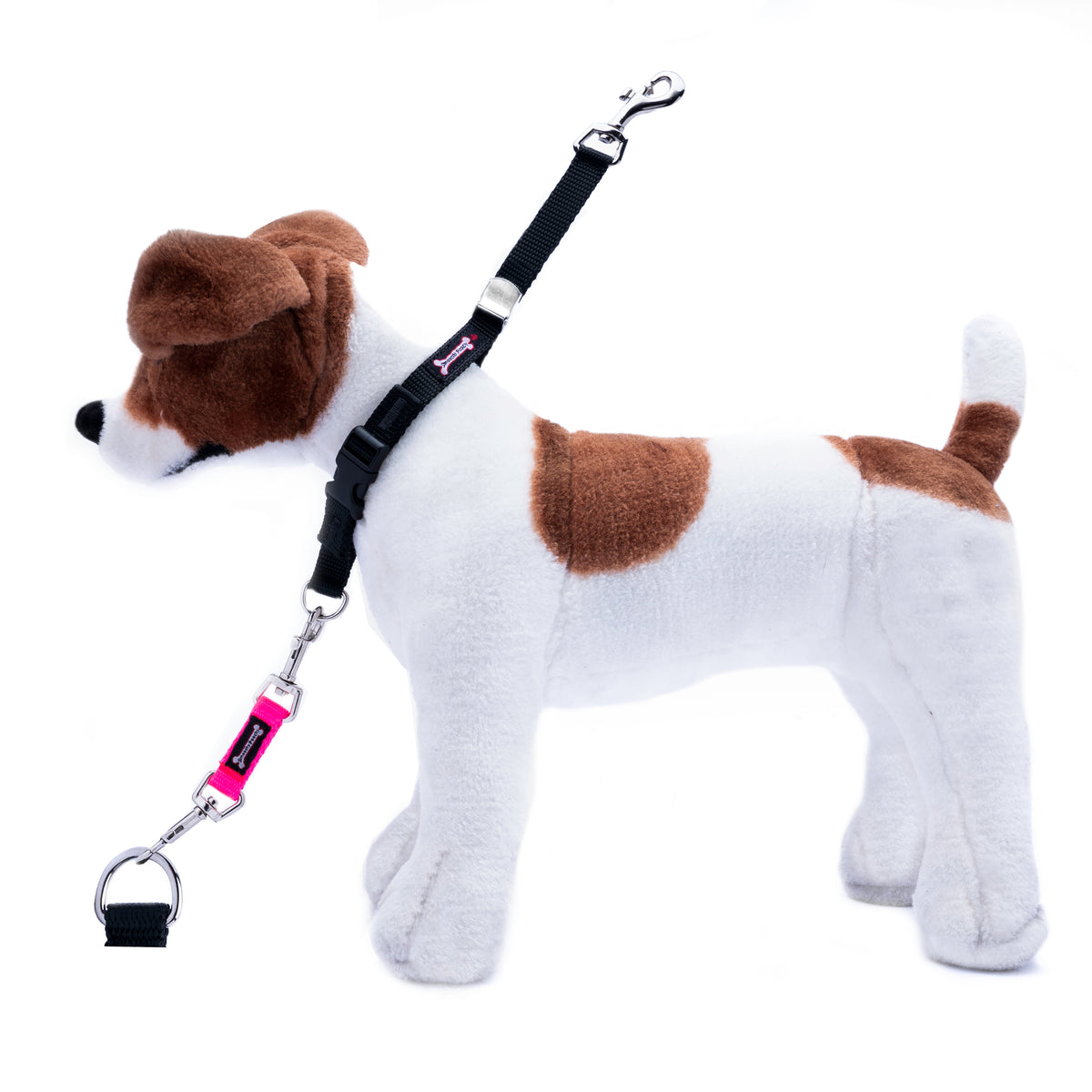 A plush toy dog in white and brown, wearing a black harness and attached pink-and-black leash